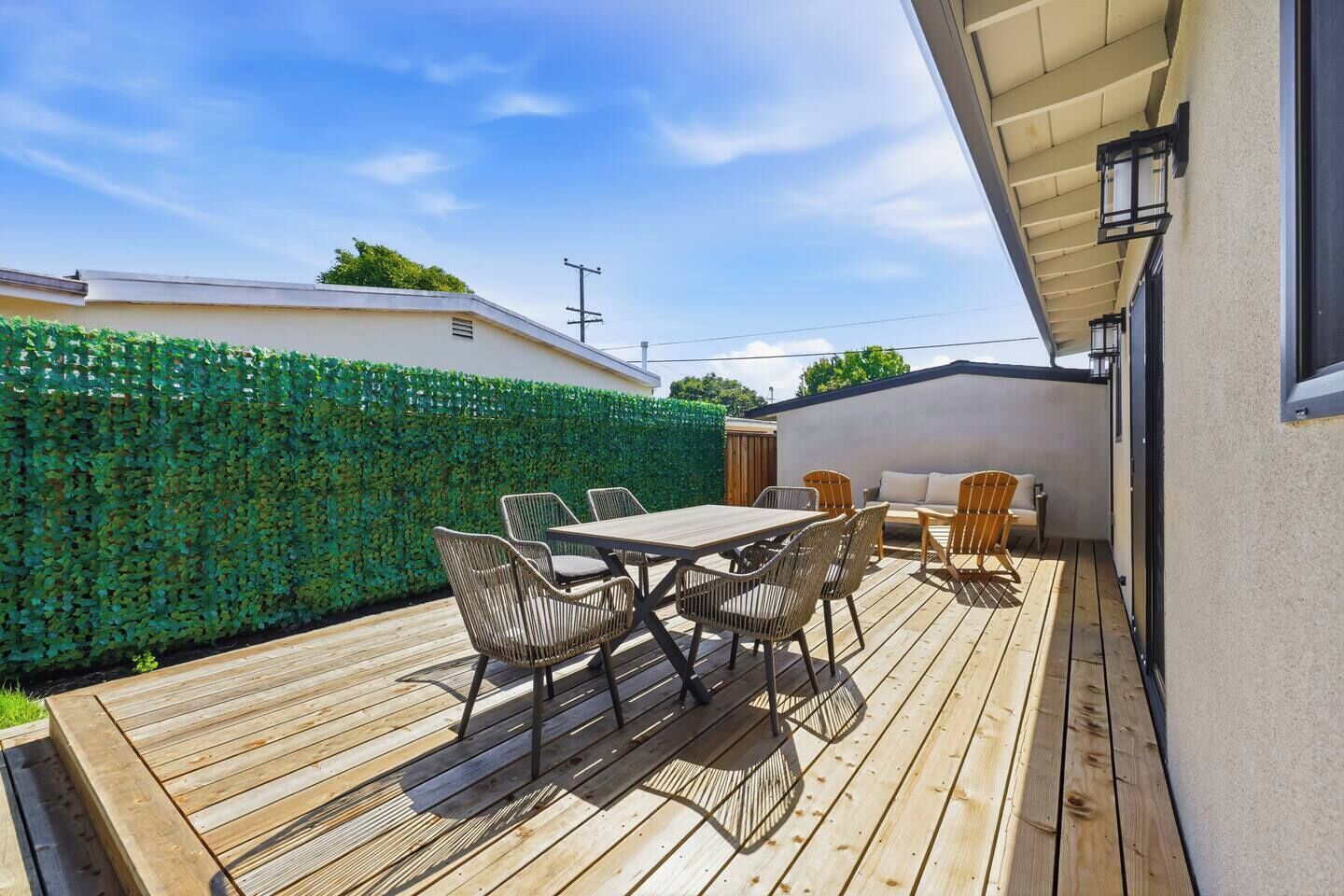 A wooden deck with a table and chairs, a green hedge, and a white building.