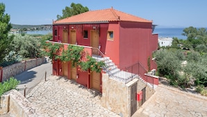 A red building with a stone staircase leading to a wooden door, surrounded by greenery and overlooking a body of water. - Classic Double Room at Hotel Aristotelis with Sea View and Balcony (Ancient Epidavros)