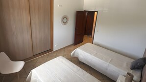 Quarto com duas camas, guarda-roupa, cadeira e mesa de cabeceira com luminária e controle remoto.