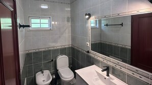 Casa de banho com lavatório branco, sanita e bidé. Há um espelho sobre o lavatório e uma janela sobre a sanita.