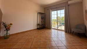 Quarto com piso de azulejos, estante, uma cadeira e uma janela grande com cortinas.