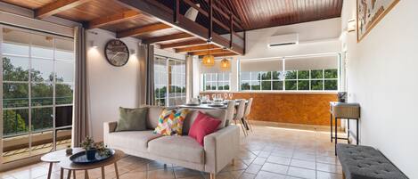 A spacious living area with a high ceiling, a sofa set, a dining table, and a view through large windows.
