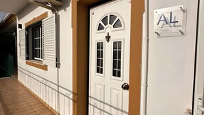 Um edifício com uma porta branca identificada com 'AL' (Alugamento Local), uma janela com persianas e um aparelho de ar condicionado fixado na parede.