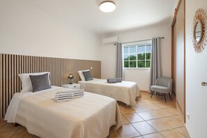 Ett hotellrum med två sängar, en stol, ett fönster med gardiner och en väggmonterad luftkonditioneringsanläggning.