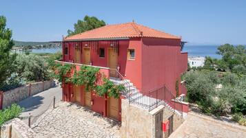 Edifício vermelho com escadaria de pedra que leva a uma porta de madeira, cercado por vegetação e com vista para um corpo d'água.