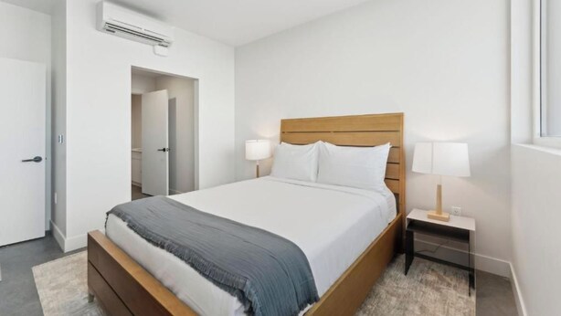 A bedroom with a wooden bed, white bedding, a nightstand with a lamp, and an air conditioning unit on the wall. - Two Bedroom Balcony with Private Hot Tub | Near City Center (Salt Lake City)