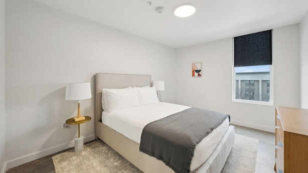 A modern bedroom with a bed, bedside lamps, a window, and a view of a building. - Modern One bedroom Unit w/ Private Laundry access in Placemakr Salt Lake City (Salt Lake City)