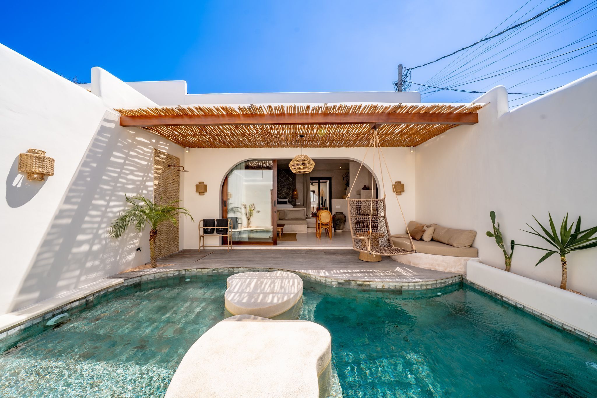 A pool with a white lounge area, a hanging chair, and a covered seating area with a view of the interior.