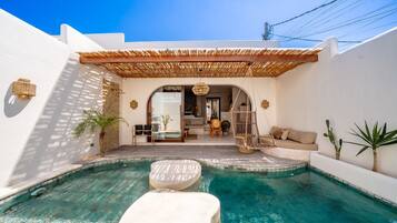 A pool with a white lounge area, a hanging chair, and a covered seating area with a view of the interior.
