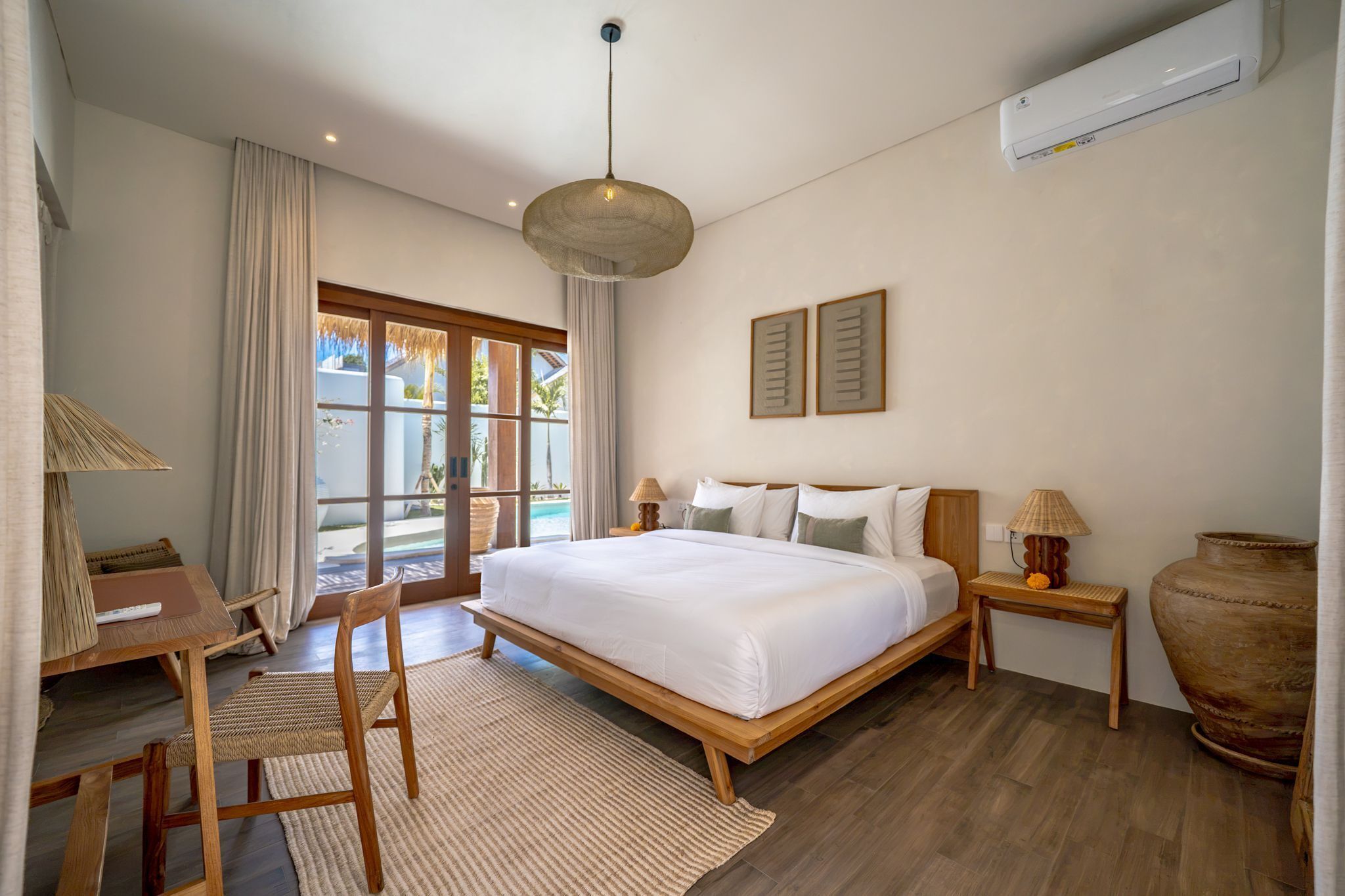 A hotel room with a bed, a desk, a chair, a lamp, a bedside table, and a large vase.