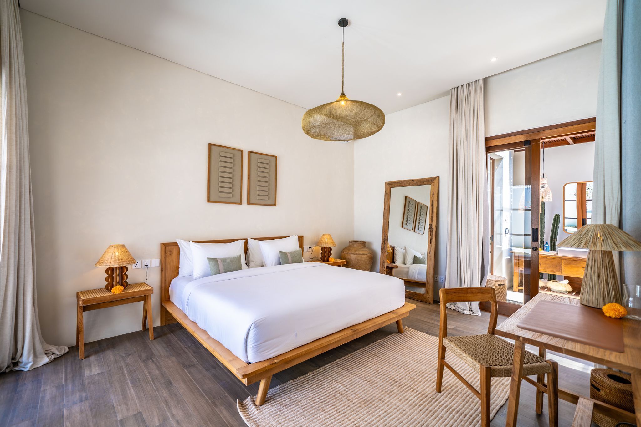 A hotel room with a wooden bed, two bedside tables, a desk with a chair, a large mirror, and a hanging light fixture.