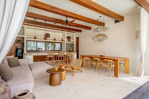 A spacious living area with a sofa, wooden dining table, and chairs, a kitchen in the background, and a ceiling fan. - Amaka Villas Uluwatu (Uluwatu)