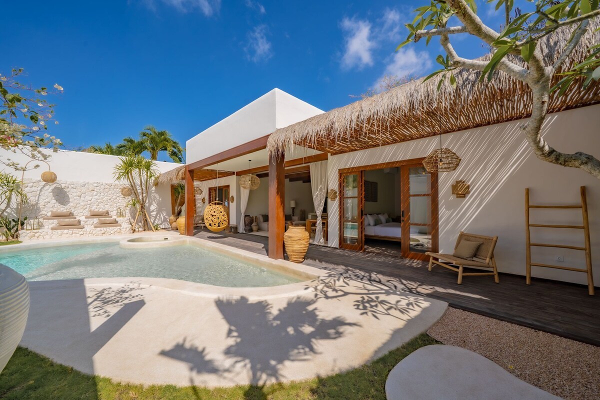 Una villa moderna con piscina, mobiliario exterior y una estructura con techo de paja.
