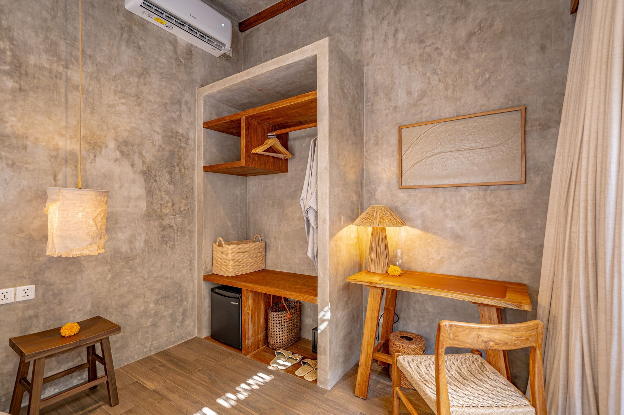 Habitación pequeña con escritorio de madera, una silla, lámpara colgante, refrigerador pequeño y aire acondicionado de pared.