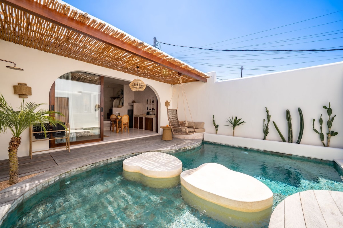 Zona de piscina con terraza de madera, una silla colgante y un comedor con puerta de vidrio.