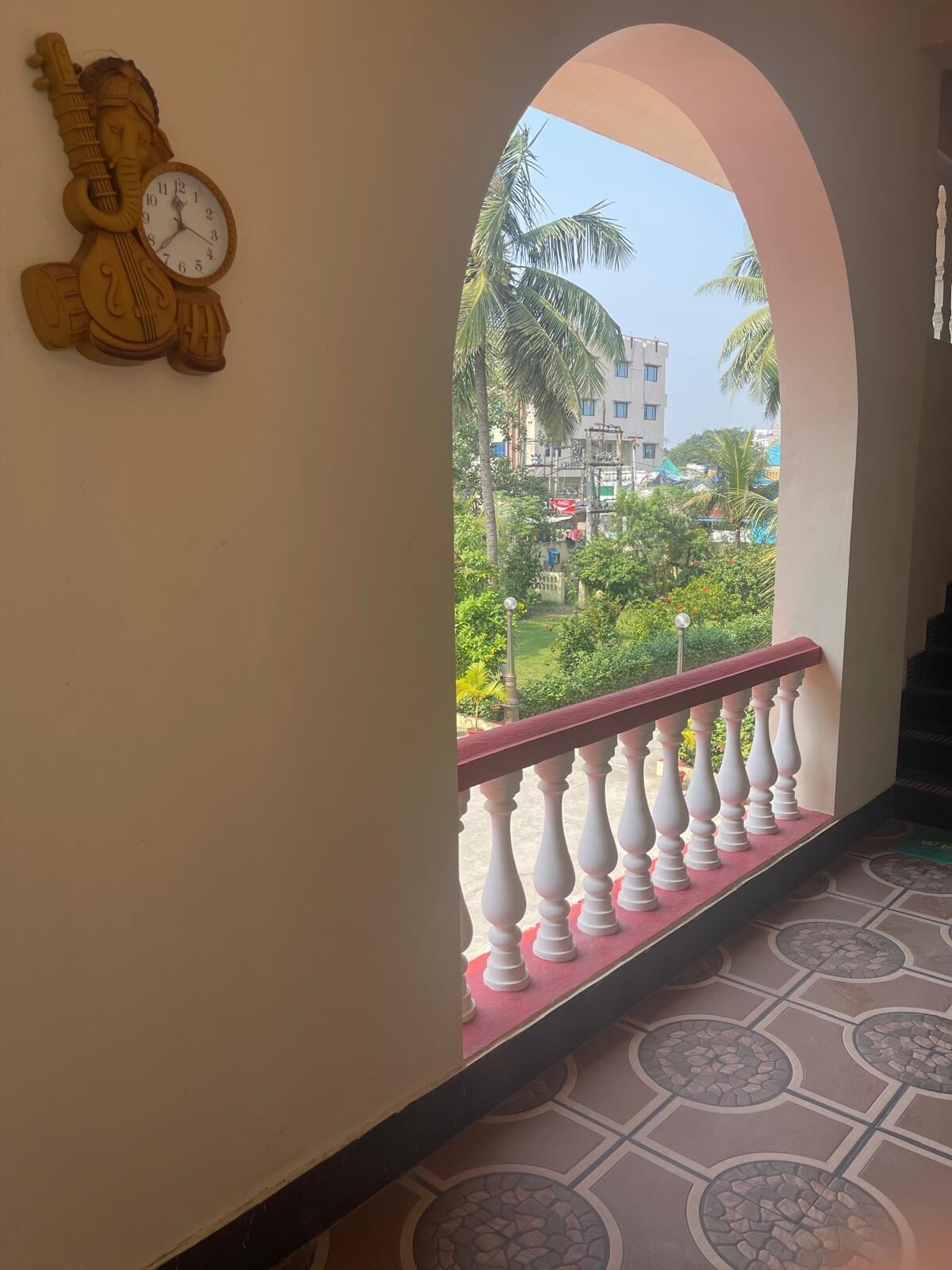A balcony with a view of a building and greenery, a clock, and a decorative wall piece.