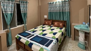 A bedroom with a bed, a ceiling fan, an air conditioning unit, a window with curtains, a bedside table, and a sink. - Sita Niwas Holiday Home Resort (Puri)