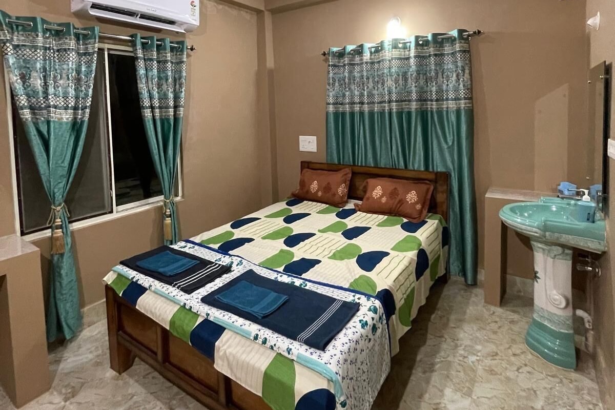 Quarto com cama, ventilador de teto, aparelho de ar condicionado, janela com cortinas, mesa de cabeceira e pia.