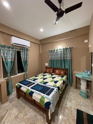 Quarto com cama, ventilador de teto, aparelho de ar condicionado, janela com cortinas, mesa de cabeceira e pia.