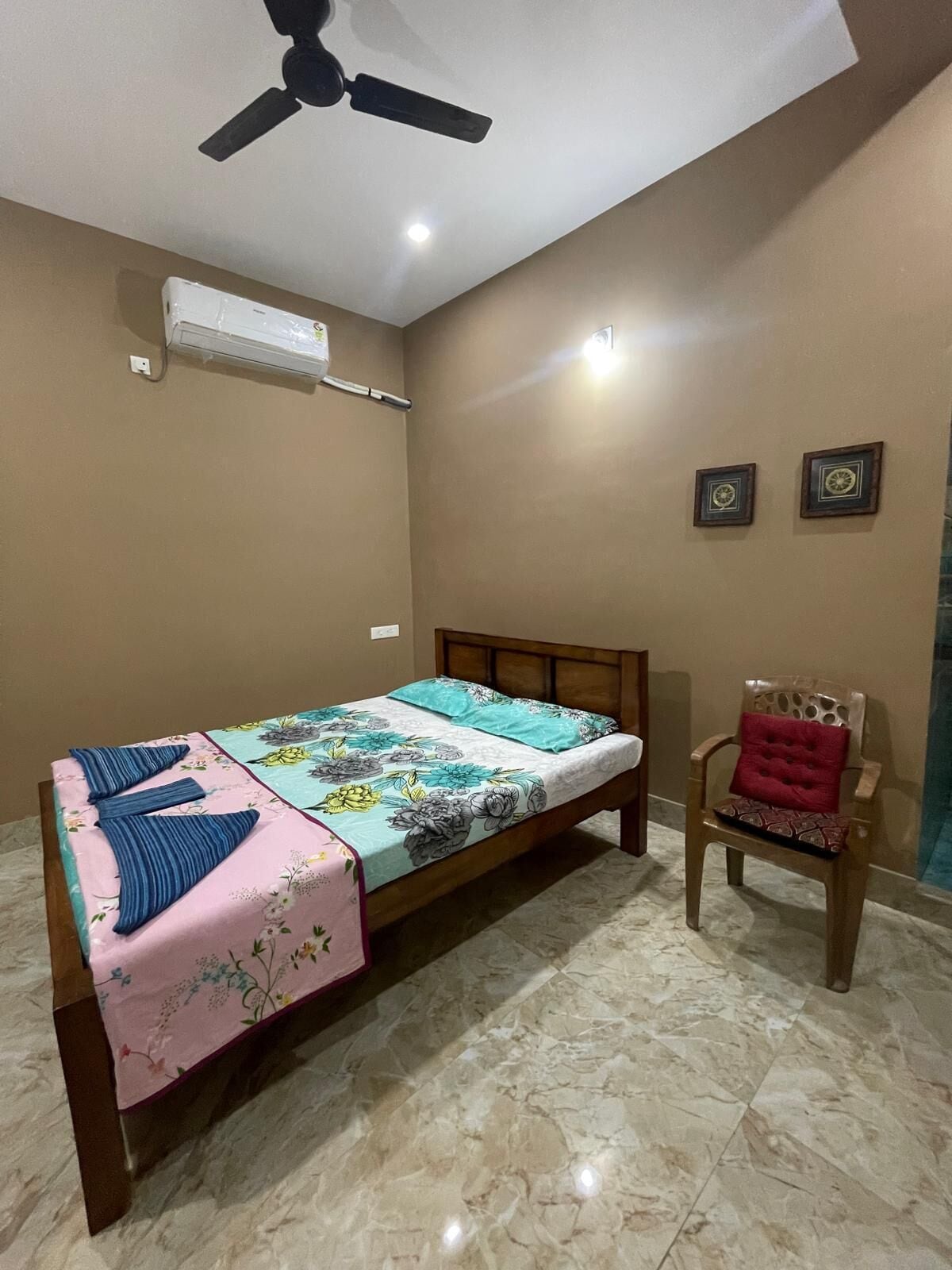A bedroom with a bed, a chair, a ceiling fan, an air conditioning unit, and two wall-mounted clocks.