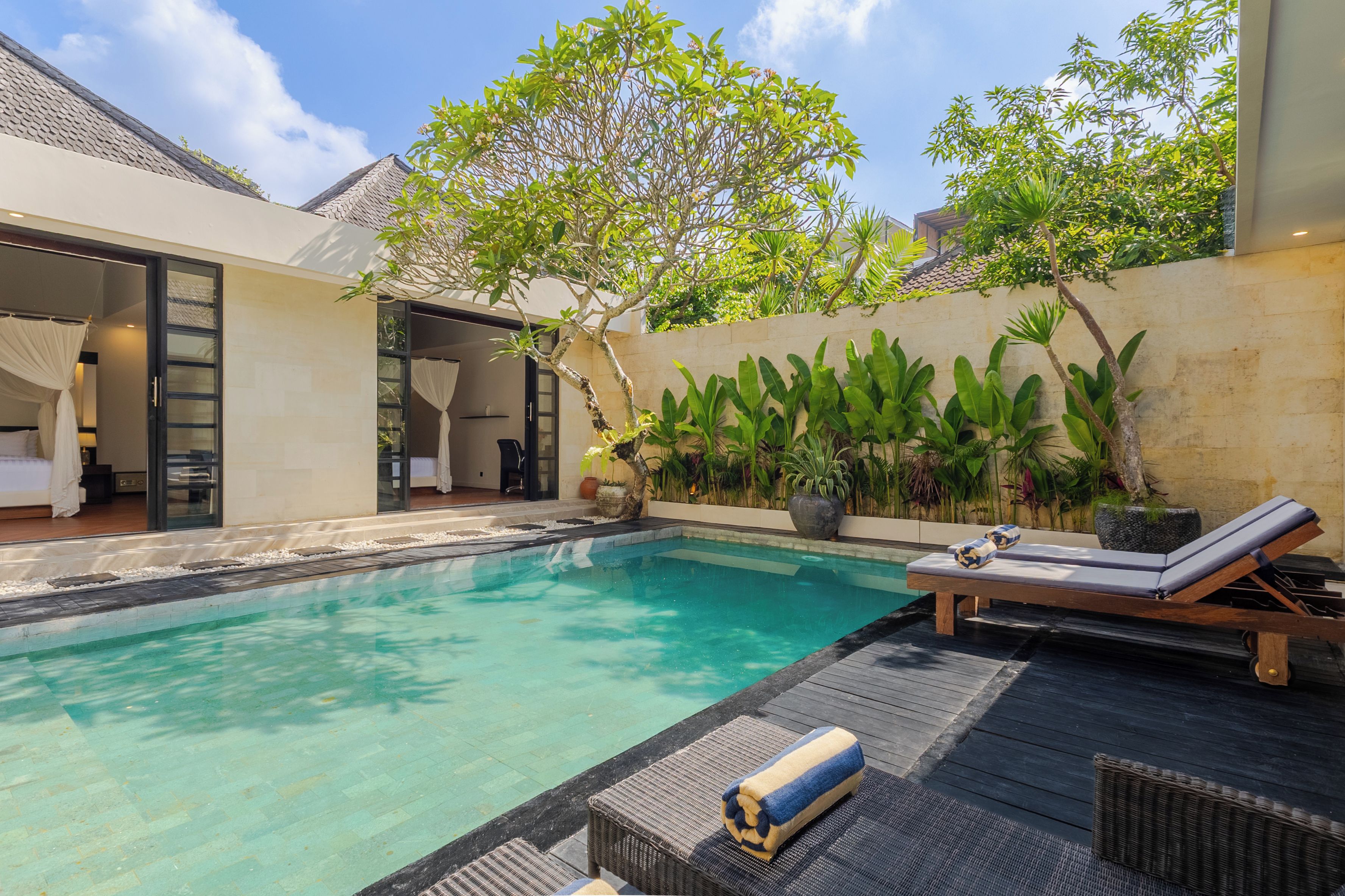 Un área de piscina privada con un sillón de descanso y una toalla, rodeada de exuberante vegetación y un edificio moderno.