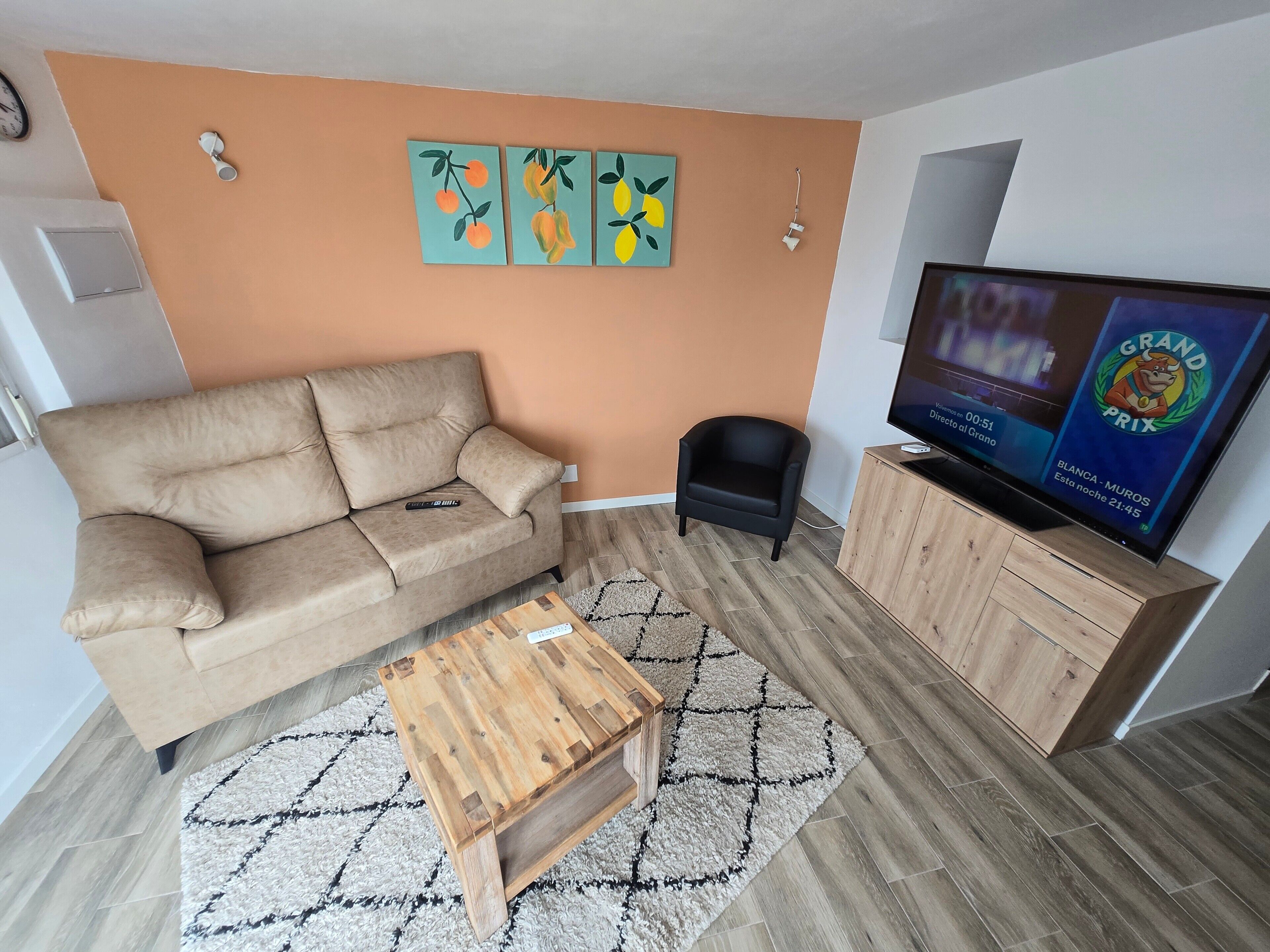 A living room with a beige sofa, a wooden coffee table, a TV on a wooden stand, and a wall with fruit paintings.