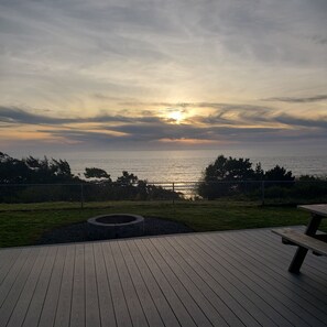 Una terraza de madera con un brasero y un banco, con vista a un cuerpo de agua al atardecer.