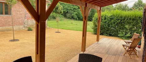A covered patio with a table and chairs, a brick building in the background, and a grassy area with trees.