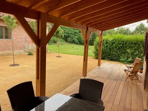 Een overdekt terras met een tafel en stoelen, een bakstenen gebouw op de achtergrond, en een gazon met bomen.