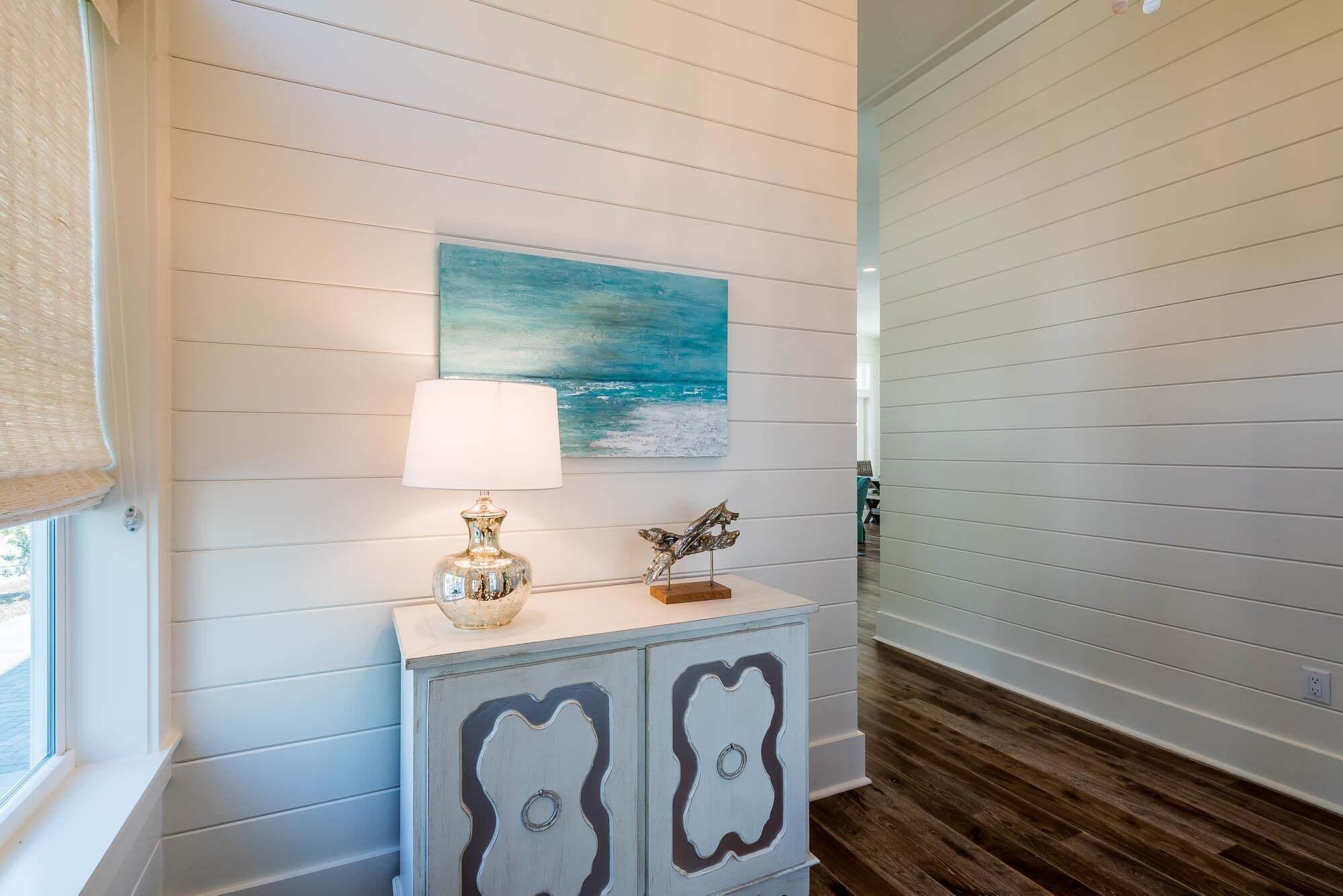 A hallway with a white wall, a painting of a blue sky and sea, a lamp, and a decorative cabinet.