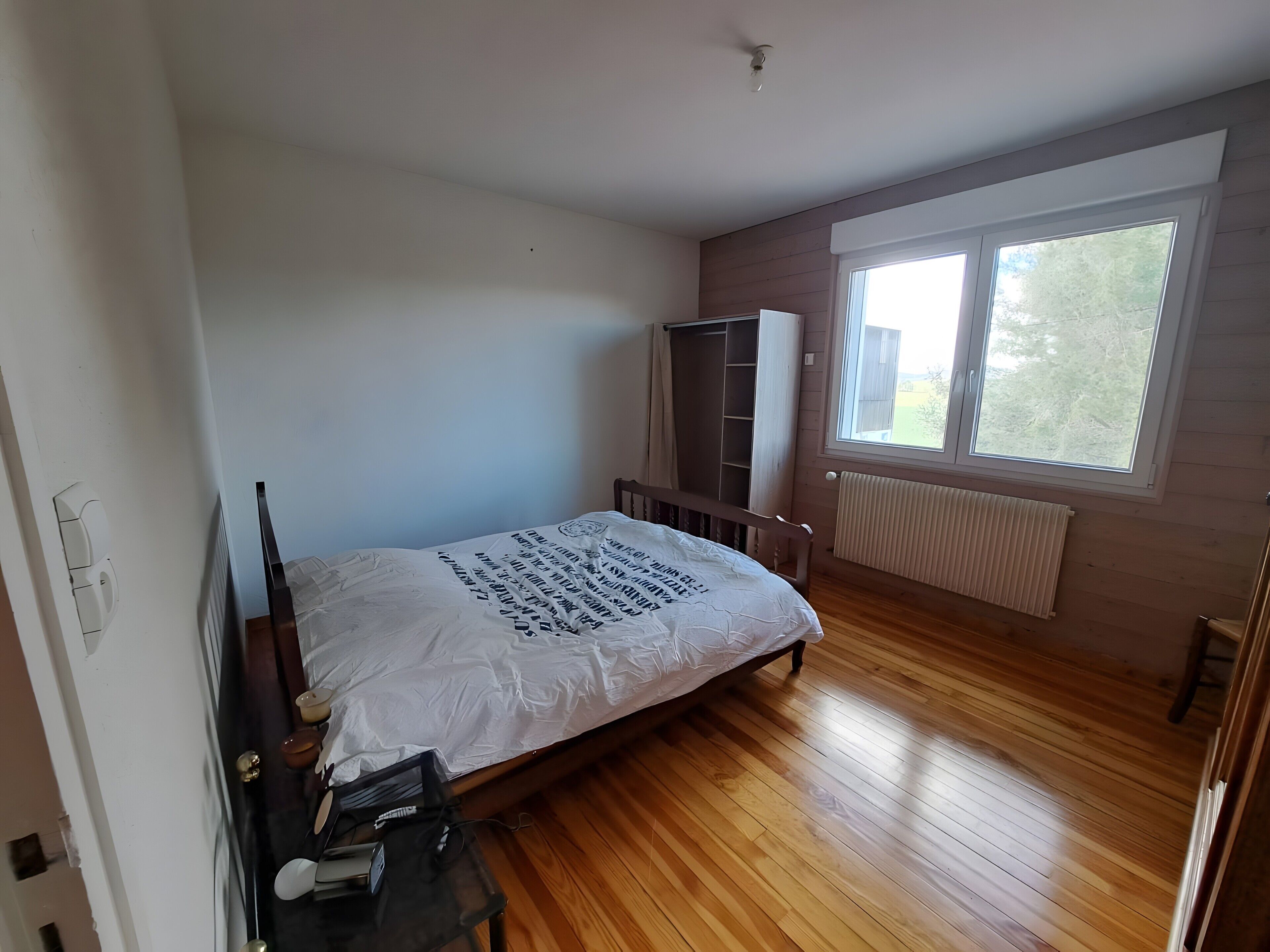 A bedroom with a wooden floor, a bed with a white mattress, a wardrobe, and a window with a view of trees.