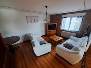 A living room with a sofa, a coffee table, a TV stand, and a window with curtains. - “Au Bonheur De La Chaux” Apartment with Private Terrace, Garden, and Wi-Fi (Damprichard)