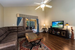 A living room with a sectional sofa, a coffee table, a TV on a wooden stand, a ceiling fan, and a patterned rug. - Our Happy Place! FREE Pool Heat (Davenport)