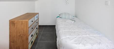 Una habitación pequeña con cama individual, un aparador de madera y una colcha blanca con estampado de hojas.