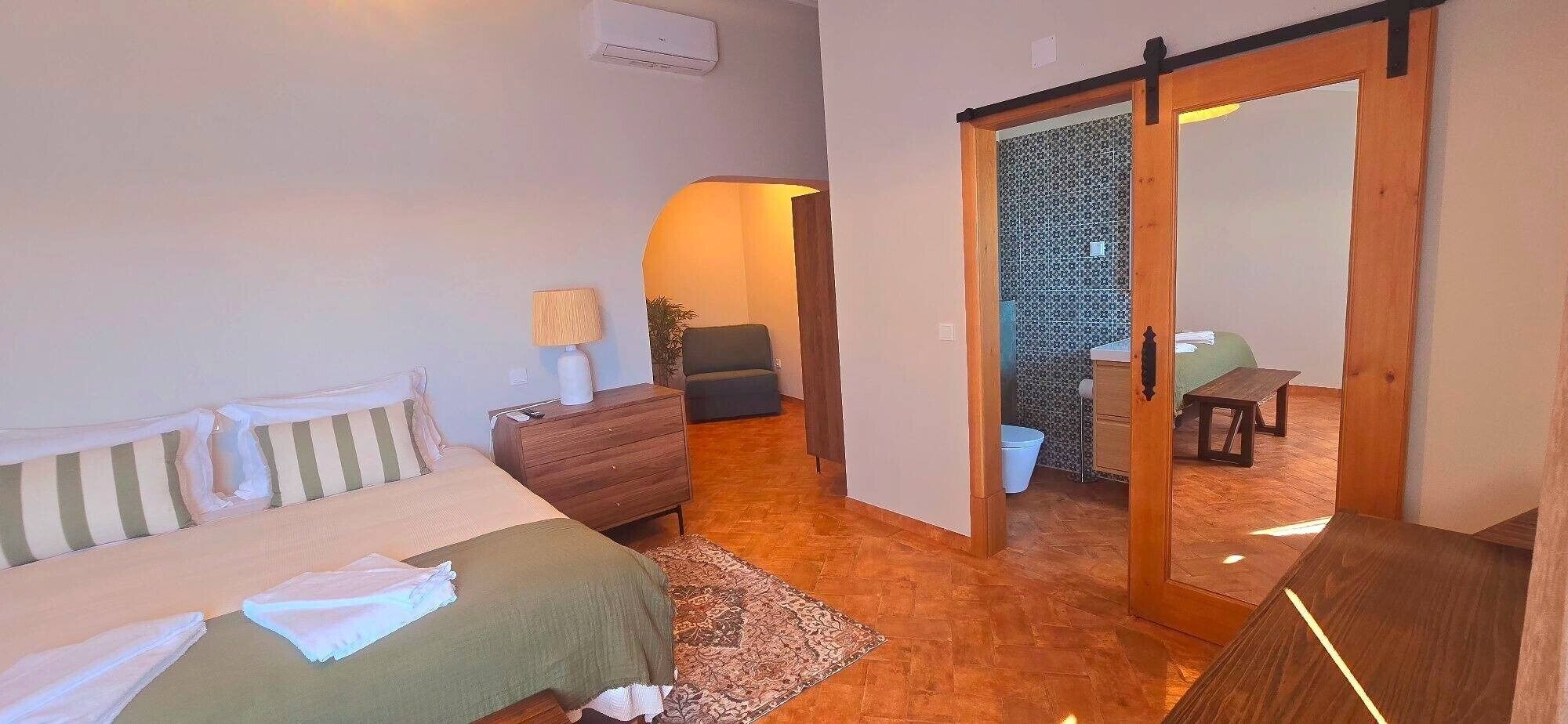 A room with a bed, a dresser, a bench, and a small table. There is a sliding door leading to a bathroom.