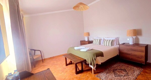 Une chambre à coucher avec un lit, des tables de chevet, un banc, un tapis et une fenêtre avec des rideaux.