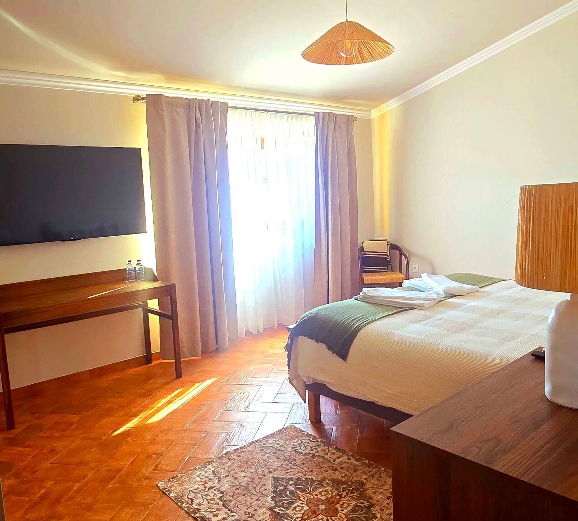 Una habitación de hotel con cama, escritorio, televisor y ventana con cortinas.