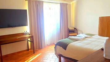 Una habitación de hotel con cama, escritorio, televisor y ventana con cortinas.