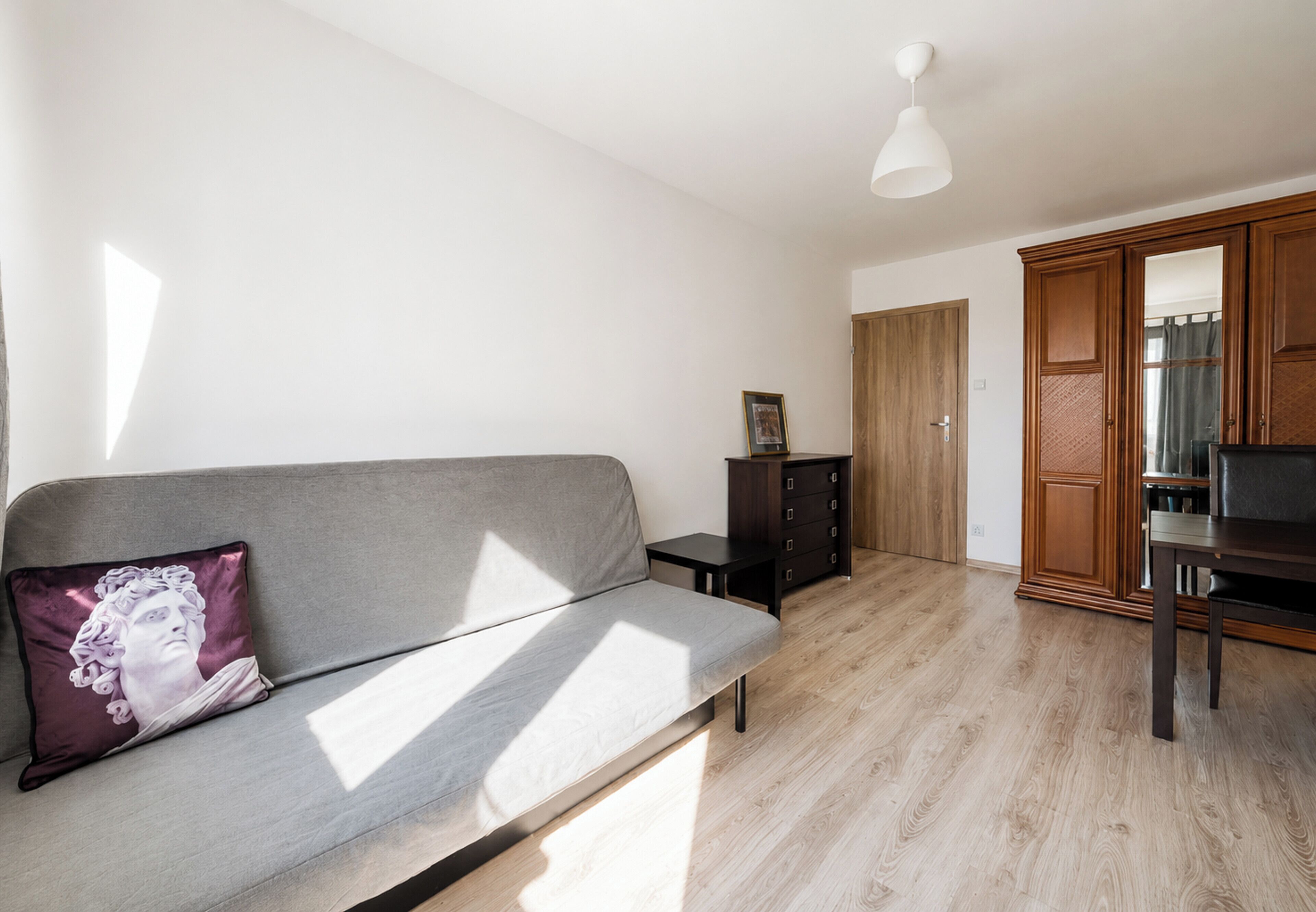 Ein modernes Wohnzimmer mit einem grauen Sofa, einem kleinen Beistelltisch, einem Holzschrank und einer Deckenleuchte.