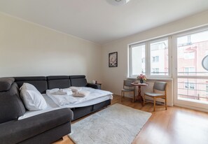 Un salon moderne avec un canapé gris, une petite table ronde accompagnée d’une chaise, et une grande fenêtre donnant sur des immeubles.