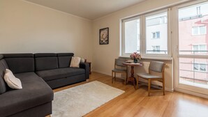 Ein Wohnzimmer mit einem dunkelgrauen Sofa, einem weißen Teppich, einem Holztisch mit Blumen in einer Vase und einem Fenster mit Blick auf Gebäude.