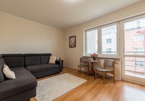 Un salon avec un canapé gris foncé, un tapis blanc, une table basse en bois ornée d’un vase de fleurs et une fenêtre donnant sur des immeubles.