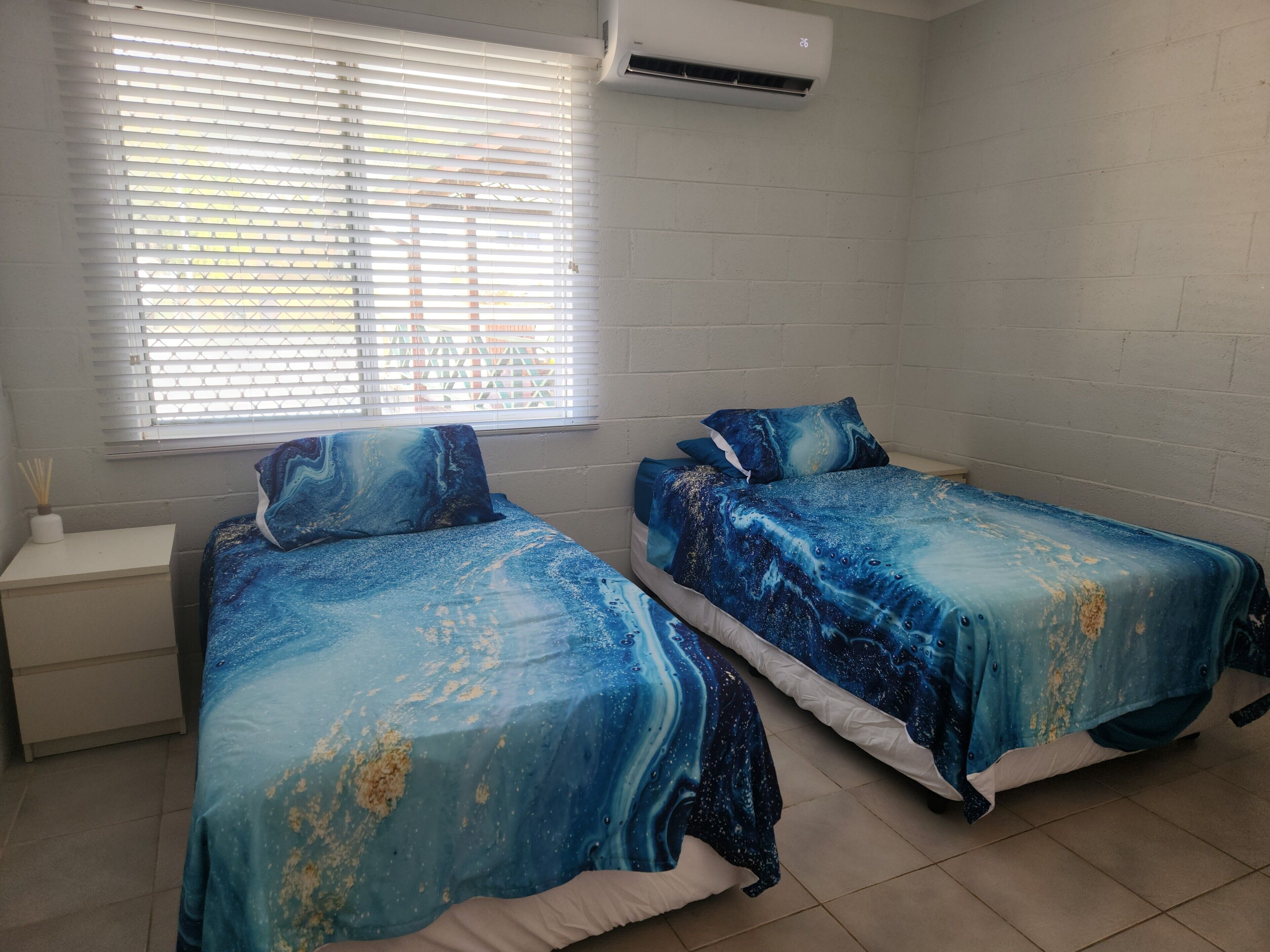 Quarto com duas camas, uma janela com persianas, ar condicionado e uma parede de tijolos brancos.