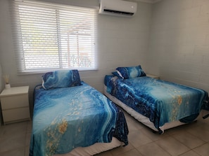 A room with two beds, a window with blinds, an air conditioner, and a white brick wall.
