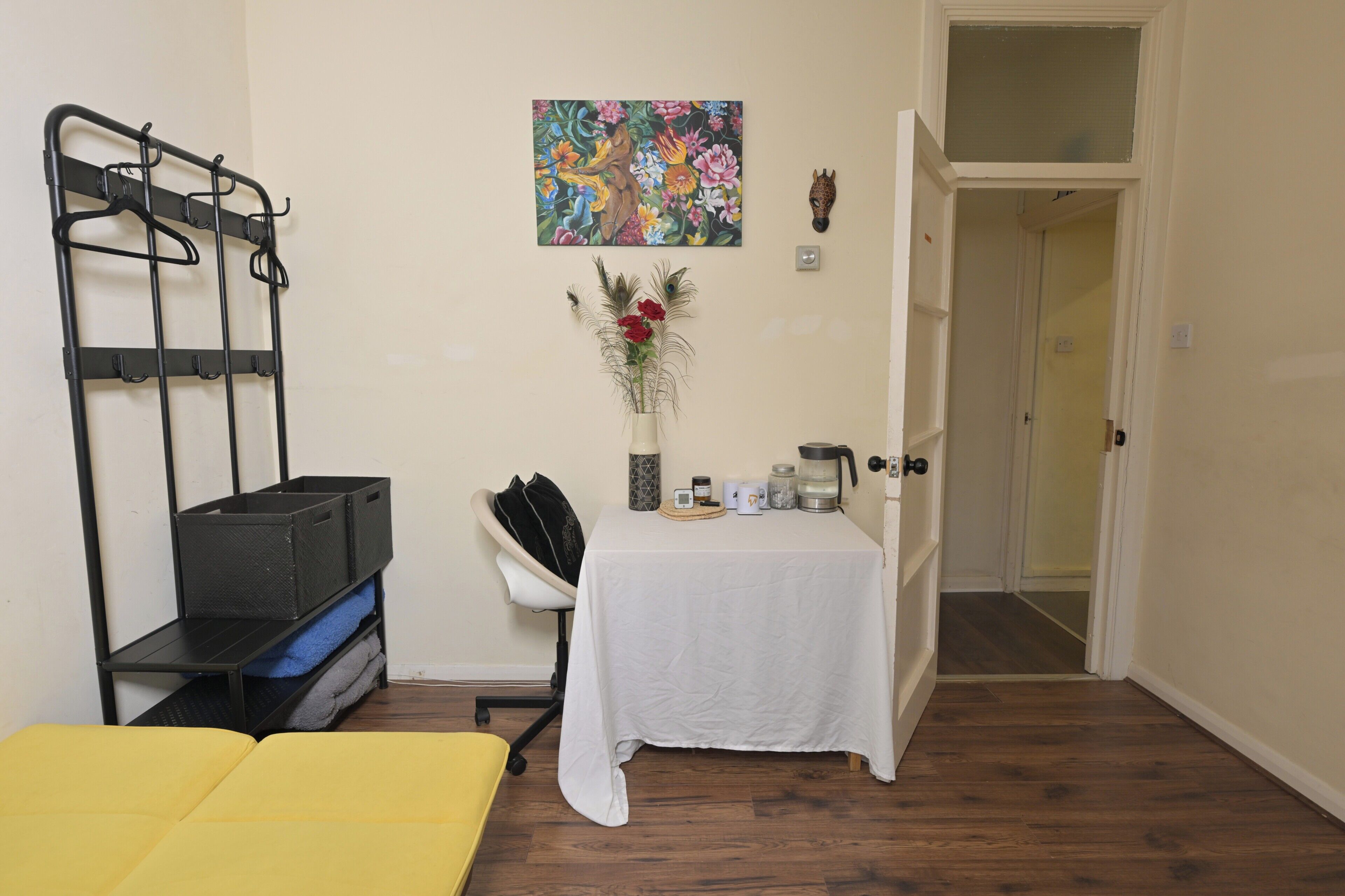 A room with a bed, a small table with items, a chair, a coat rack, and a door leading to another room.