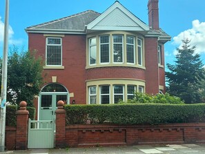 A two-story house with a bay window, a front door with a green gate, and a brick wall with a hedge. - 3 bedroom Apartment 2 up to 5 people Free parking (Blackpool)
