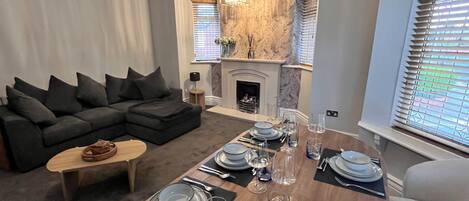 A modern living room with a sectional sofa, a dining table set for four, and a fireplace.