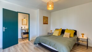 Quarto com cama, mesas de cabeceira, luminária pendente, porta de acesso a um banheiro e um quadro na parede.