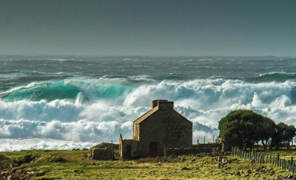 A coastal scene with a stone building, waves crashing, and a fenced area with trees.