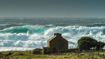 A coastal scene with a stone building, waves crashing, and a fenced area with trees.